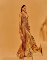 Vestido largo de tejido crochet en color bronce, con escote en V profundo, diseño de red y detalles trenzados. Presenta una silueta ajustada y un acabado con flecos largos en el ruedo.