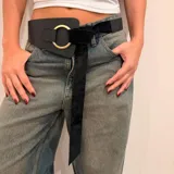 Cinto ancho de cuero marrón con diseño asimétrico, que presenta un aro metálico dorado como elemento central y una tira de gamuza larga que se anuda para el cierre.