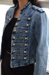 Chaqueta de estilo militar confeccionada en denim negro con efecto lavado. Presenta un diseño estructurado con cierre frontal de cremallera, cuello mao y múltiples botones metálicos decorativos dispuestos en filas horizontales. Incluye detalles de charreteras en los hombros y puños con botones.