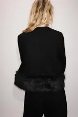 Chaqueta de punto negra con cuello redondo y manga larga, con detalles de pelo sintético a tono en los puños y en el bajo. Cuenta con cierre frontal con botones a presión ocultos.