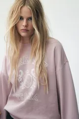 Buzo de tejido de punto color rosado pálido, con cuello redondo, hombros caídos y puños y bajo acanalados. Presenta un estampado frontal en blanco con el texto "Firenze Italia" y un escudo central con el año "1932".