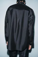 Camisa de corte holgado confeccionada en mezcla de lyocell con acabado satinado. Presenta cuello redondo con lazadas integradas, cierre frontal con botones ocultos bajo solapa, bolsillos de plastrón en el frente y mangas largas con puños anchos. El bajo es asimétrico y redondeado.