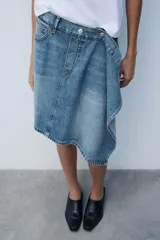 Falda de tiro medio confeccionada en denim, con trabillas, cinco bolsillos, cierre cruzado con botones y bajo acabado asimétrico.