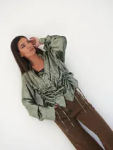 Camisa verde con textura brillante, cuello camisero, mangas largas con puños abotonados y cierre frontal con botones de nácar. Presenta frunces con cordones ajustables en la parte delantera.