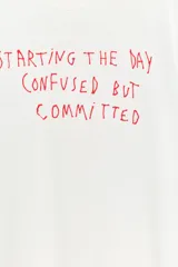 Camiseta blanca de algodón flamé, con cuello redondo, manga corta y estampado frontal de texto en color rojo con la frase 'Starting the day confused but committed'.