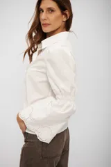Camisa blanca de corte clásico con cuello camisero y cierre frontal de botones. Destaca por sus mangas largas con detalle de frunce a la altura del codo y terminación acampanada con bordado inglés.