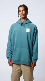 Hoodie de color verde azulado con capucha ajustable mediante cordones. Presenta un bolsillo frontal tipo canguro y un parche cuadrado en el pecho con una cruz blanca sobre fondo amarillo.