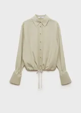 Camisa color beige de corte crop, confeccionada en mezcla de lino y lyocell. Tiene cuello camisero, mangas largas con puños abotonados, lazo ajustable en la cintura y cierre frontal con botones.