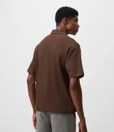 Remera polo de manga corta y corte regular, confeccionada en tejido texturizado color marrón, con cuello camisero, cartera corta con botones y un bolsillo aplicado en el pecho.