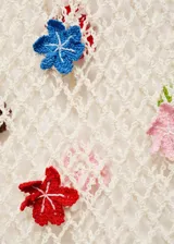 Vestido largo de tejido calado en color crudo, con diseño de red y aplicaciones de flores tejidas en diversos colores. Presenta escote en V con lazo, manga larga y corte entallado.