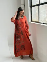 Vestido largo color terracota con diseño asimétrico que incluye un lateral con estampado floral grande. Presenta mangas tres cuartos, escote redondo y un bolsillo aplicado en el frente.
