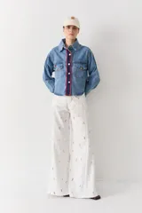 Campera de denim azul con corte clásico, cierre frontal con botones metálicos dorados y detalle de tejido crochet en contraste en color azul marino y rojo a lo largo de la solapa. Presenta bolsillos frontales con solapa y botones.