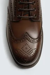 Zapato tipo derby de hombre, color marrón oscuro, fabricado en piel con detalles de picados (brogue) en puntera y laterales. Posee plantilla interior de piel y suela track con ligero tacón.