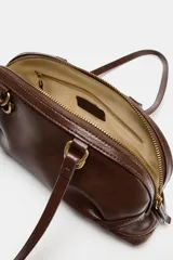 Bolso de hombro de color marrón, con diseño alargado y detalle de anillas metálicas doradas en la base de las asas. Cuenta con cierre de cremallera superior y doble asa fina para llevar colgado.