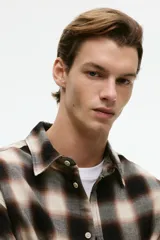 Camisa de manga larga confeccionada en franela de algodón suave, con diseño de cuadros en tonos marrones y beige. Presenta cuello inglés, cierre frontal con botones y bajo redondeado.
