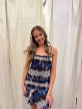 Vestido corto de tirantes finos con estampado tie-dye en tonos azul, negro y gris. Presenta un escote recto con frunce central y un volado en el ruedo de una textura diferente.