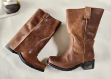 Bota de cuero marrón con diseño 2 en 1, que incluye una polaina desmontable. Presenta una caña alta estructurada y una base de bota tipo botineta con suela plana de goma negra.