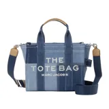 Bolso tipo tote de Marc Jacobs, modelo Mini Traveler, confeccionado en denim con diseño de patchwork en diferentes tonos de azul. Presenta el logo de la marca estampado en el frente, doble asa superior y correa de hombro ajustable y extraíble en color azul marino con detalles en cuero tostado.