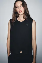 Chaleco negro de cuello redondo y sin mangas, con cierre frontal cruzado con un botón y bajo asimétrico.