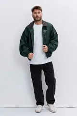 Cazadora de estilo urbano en color verde oscuro, con cuello de pana en tono marrón, cierre frontal con cremallera y puños elásticos. Presenta un diseño holgado con bordado pequeño en el pecho.