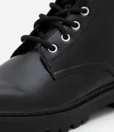 Bota estilo coturno de caña media, confeccionada en poliuretano con acabado liso. Presenta cierre mediante cordones frontales con ojalillos metálicos y suela track robusta.