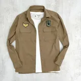 Sobrecamisa color beige de gabardina, con cuello clásico, cierre frontal con botones, dos bolsillos de parche con solapa abotonada en el pecho y parches bordados en el pecho y el hombro.