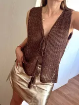 Chaleco tejido en punto calado con hilo de algodón y lurex, de color negro con destellos plateados. Presenta escote profundo en V, cierre frontal con cordones entrelazados terminados en borlas y un corte recto y holgado.