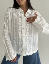Camisa blanca de manga larga con delicados bordados florales verticales y detalles de encaje. Presenta cuello clásico, cierre frontal con botones y terminaciones festoneadas en los puños y el ruedo.