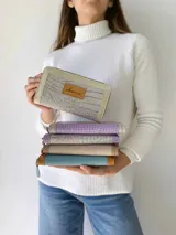 Billetera grande con cierre metálico, color beige y crema. El frente presenta un tejido texturado con patrón de rayas horizontales en tonos claros, ribetes en charol color crema y un parche de cuero marrón con la marca grabada.