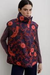 Chaleco puffer con cuello alto y cierre frontal oculto, estampado con diseño floral y de aves en tonos rojos sobre base azul oscuro.