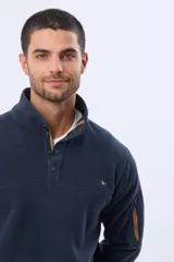 Buzo de abrigo confeccionado en tejido polar térmico, diseñado para mantener el calor y ofrecer resistencia al viento. Presenta un cuello alto con cierre frontal de broches, bolsillos laterales con cierre y un bolsillo adicional con cierre en la manga izquierda.