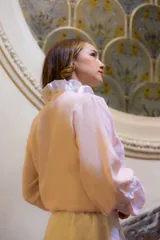 Camisa de lino color rosa pálido, con cuello alto decorado con volados, cierre frontal con botones forrados y mangas largas amplias con puños terminados en volados.