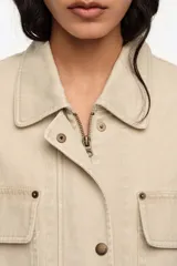 Chaquetón de corte recto color beige, confeccionado en algodón, con cierre frontal de cremallera oculta y botones a presión. Posee cuello camisero, mangas largas, bolsillos con solapa en el pecho, bolsillos tipo plastrón en la parte inferior y un pequeño bolsillo con cierre en la manga.