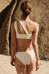 Top de bikini asimétrico de un solo hombro en color beige, con doble tirante y un detalle de aplique metálico dorado en la parte delantera.