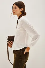 Camisa blanca con cuello mao y escote en V, con bordados negros y dorados en el frente y mangas largas con puños abotonados.