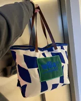Bolso tote de tela color blanco con estampado de manchas azules. Tiene asas de cuero marrón y un bolsillo frontal con parche verde bordado con el logo de Willa Squad en azul.