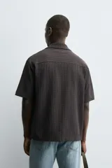 Camisa de manga corta con cuello bowling y calce relaxed fit, confeccionada en tejido seersucker elástico con textura. Presenta un diseño a rayas verticales en tonos oscuros y cierre frontal con botones.