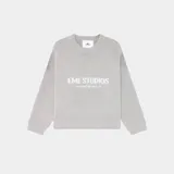 Sweater de tejido de punto, color gris claro, con cuello redondo y manga larga. Presenta un estampado frontal con el texto "EME STUDIOS" y un texto más pequeño debajo.