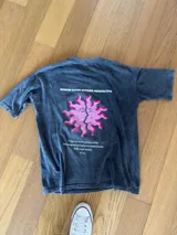 Remera de algodón con efecto lavado, corte holgado y diseño gráfico en la espalda que presenta un sol con rostros en color rosa y texto impreso.