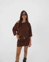Vestido corto color marrón, de corte holgado y diseño minimalista con escote asimétrico que deja un hombro al descubierto y mangas tres cuartos.