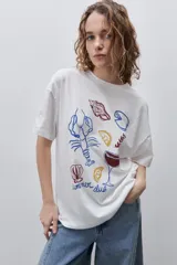 Remera blanca de silueta amplia y calce relajado, con cuello redondo, mangas cortas con hombros caídos y estampa gráfica multicolor en el frente con motivos de verano.