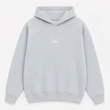 Canguro con capucha de calce oversize, confeccionado en tejido pesado color gris claro. Presenta un bordado minimalista con la palabra 'ASCEND' en la parte superior trasera, puños y ruedo acanalados.