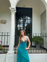 Vestido largo con estampado tie-dye en tonos violeta y fucsia, escote en V con detalles bordados dorados, cut-outs laterales bajo el busto y tirantes finos que se cruzan y atan en la espalda.