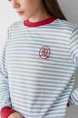 Remera de manga larga con diseño de rayas horizontales en color celeste y blanco. Presenta cuello redondo y puños en color rojo contrastante, junto con un pequeño parche bordado en el pecho.