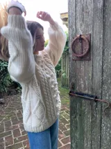 Sweater artesanal de tejido de punto de lana merino color marfil, de calce amplio y relajado, con cuello redondo y diseño frontal en relieve con trenzas y bodoques. Presenta mangas largas abullonadas con paneles texturizados y puños acanalados.