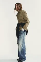 Chaqueta de estilo militar en color beige, con cuello subido, cierre frontal de botones, bolsillos de plastrón con solapa en el delantero y detalle de solapas en el pecho. Presenta mangas largas con hombreras y trabillas decorativas.