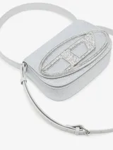 Bolso bandolera Diesel modelo 1DR, color plateado con glitter, con asa corta y correa larga extraíble. Presenta solapa con cierre magnético y logo ovalado "D" metálico en el frente.