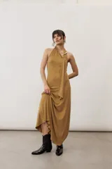 Vestido largo color tostado de corte fluido, con escote halter y detalle de tiras cruzadas con aplique metálico en el hombro.