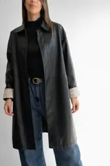 Trench de ecocuero negro de corte relajado y largo medio. Presenta cierre frontal con botones, bolsillos delanteros funcionales y tajo posterior. Las mangas cuentan con un forro interno a rayas que queda visible al doblarlas.