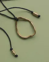 Collar con dije colgante de forma orgánica e irregular, acabado dorado, suspendido de un cordón negro de algodón natural.